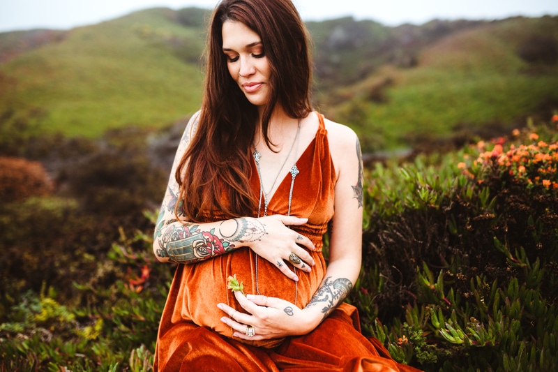 Maternity Photography, An expectant mother sits in grassy hills holding her belly