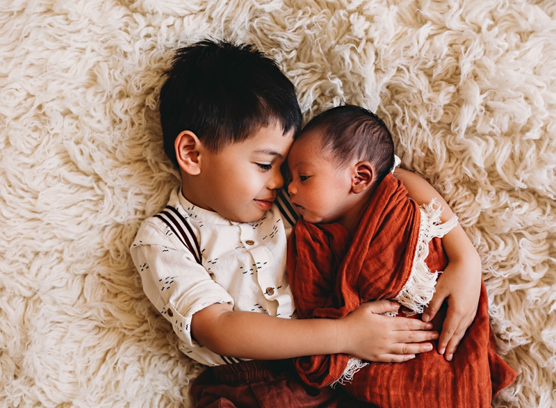 Newborn Photography, a newborn baby sleeps swaddled in a blanket, older brother holds baby close