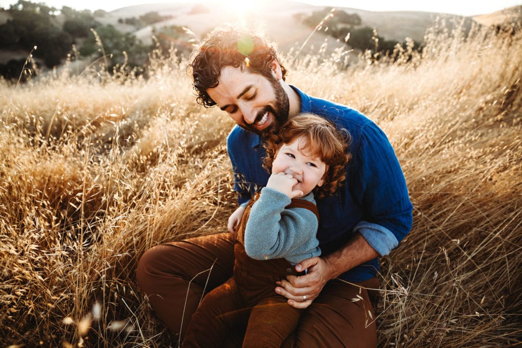 Family Photographer, a father admires his toddler son, the boy sits on his lap outdoors on a dry grassy hillside